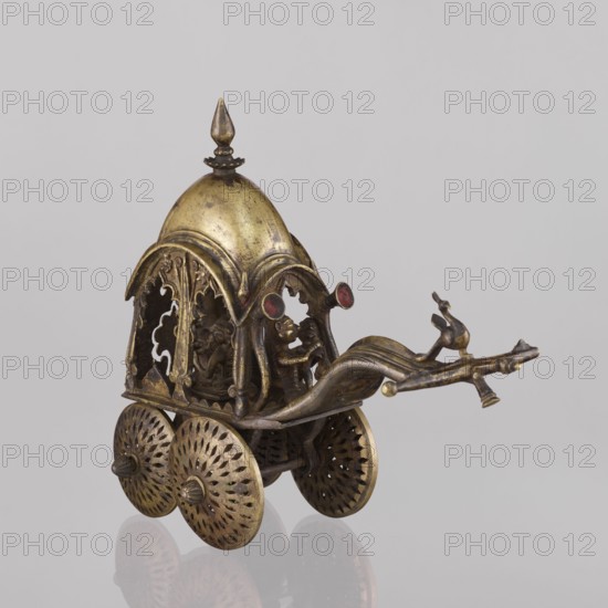 Miniature of a chariot, chariot model, 19th century, India; Indian; South Asian; temple chariots; made by, height 24.0 cm, width 28.0 cm. A bird sits on a bow-shaped shaft; peacocks and other birds depicted. Under a canopy topped with a pike sit figures: Krishna feeding on butter; in front, in the driver's seat, there is also Krishna., A small ornate metallic chariot-like object in gold and brass tones sits against a pale gray background, featuring a domed canopy with a pointed finial, arched openwork sides with decorative cutouts, a curved front extension with a sculpted animal figure, and a few small stylized human figures visible within the open cabin; the piece rests on a group of round, perforated wheels with radial patterns and shows areas of darker patina and subtle reddish accents.