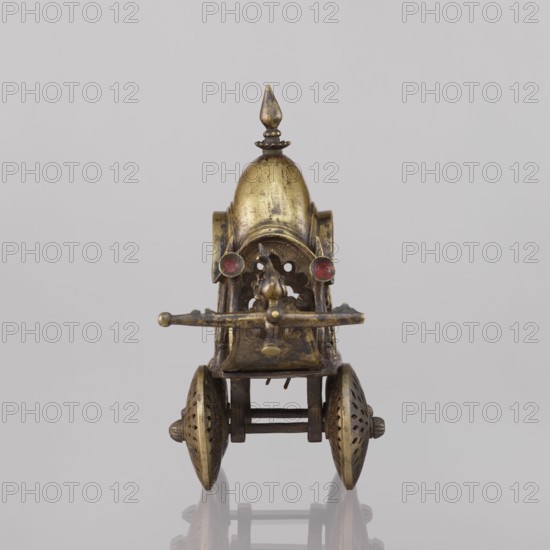 Miniature of a chariot;, 19th century, India; model; height 24.0 cm, width 28.0 cm. Temple chariot motif with peacocks and birds. A bird sits on a bow-shaped shaft. Under a canopy topped with a pike sit figures: Krishna feeding on butter; in front, in the driver's seat, there is also Krishna. Indian art; South Asian., A small metallic wheeled object viewed from the front, with a domed top and a pointed finial, showing a worn brass or gold-toned surface with darker patina; a pair of round red accents sit near the front of the dome; a horizontal bar protrudes across the front above an open lattice-like opening with curved decorative cutouts; two ornate spoked wheels with perforated patterns flank the lower sides, connected by a rectangular axle structure in a darker metal; the background is a smooth, light gray surface and the object casts a faint reflection below.
