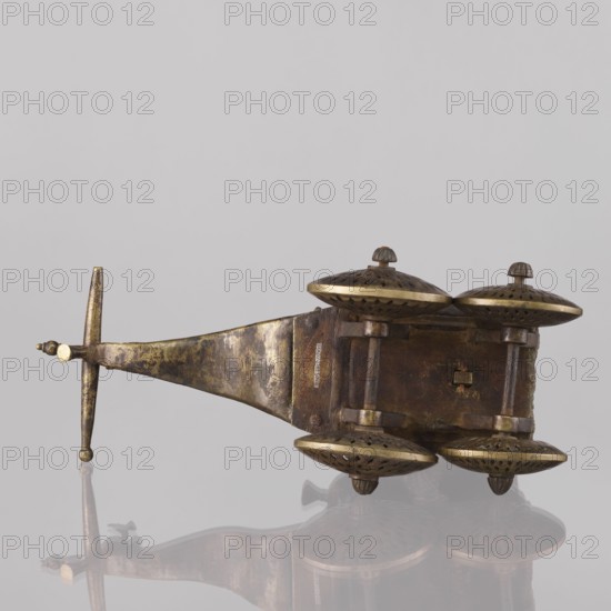 Miniature of a chariot; maker, 19th century India; model of a temple chariot depicting peacocks and birds. A bird sits on a bow-shaped shaft. Under a canopy topped with a pike sit figures: Krishna feeding on butter; in front, in the driver's seat, there is also Krishna. Materials/technique not specified. Height 24.0 cm, width 28.0 cm., An aged metallic object with a long tapered handle and a crossbar end lies on a reflective surface; the metal shows a mottled patina of brown, dark brown, and muted gold tones. Attached to the wider end of the object is a rectangular block from which a group of dome-shaped caps project; the domes are bronze-colored with darker perforated patterns and lighter golden rims, and each dome has a small rounded finial on top. The rectangular block and connecting posts share the same brown and gold worn metallic finish, and the reflection beneath the object repeats the brown, gold, and darker shadowed hues. The background is a smooth pale gray.