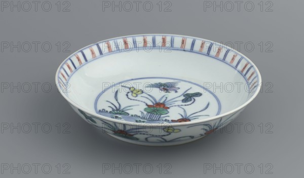 Plate, Chinese art, Chinese, Qing dynasty 1644–1911, ceramics—colored porcelain decorated with lotuses and ducks, decoration in mirror and outside, 1st half of the 18th century, China, vessel, height 3.5 cm, diameter 15.8 cm, A shallow round ceramic bowl with a white base color, decorated inside and along the rim with painted floral and leaf motifs in blue, green, red, and yellow; the rim also shows a repeated vertical band pattern in blue and red. The interior central area features a circular border in blue surrounding stylized plants and flowers in blue, green, red, and yellow. The exterior side of the bowl displays a continuous floral design in the same blue, green, red, and yellow palette. The bowl sits on a smooth gray surface against a plain gray background.