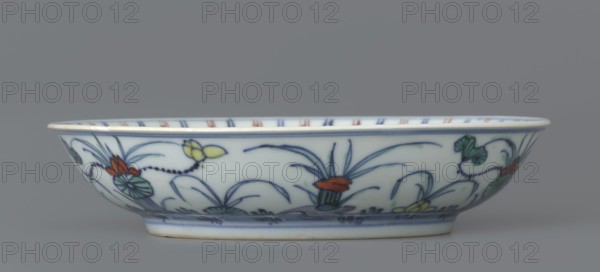 Plate, gift, Chinese, Qing dynasty 1644–1911, colored porcelain, decorated with lotuses and ducks in mirror and outside, 1st half of the 18th century, China; vessel; height 3.5 cm, diameter 15.8 cm, A shallow oval ceramic bowl with a white base color sits against a gray background, decorated around its exterior with hand-painted floral motifs in blue, green, red, yellow, and black, featuring stylized leaves, petals, and curved stems; the rim has a thin blue band and the interior rim shows alternating short vertical stripes in blue and red, while the underside near the foot has a blue horizontal band and the foot rim shows a slightly warm off-white tone.