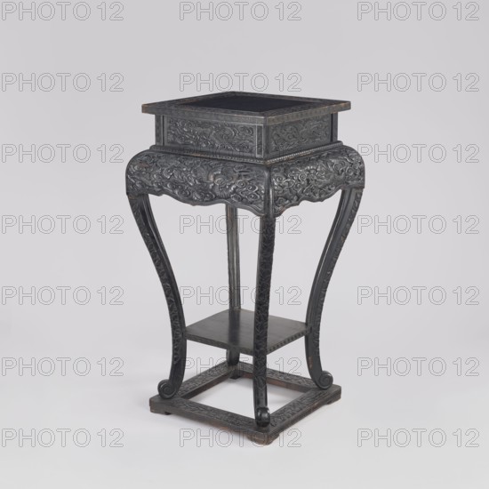 Table, phoenixes iconography, dragons iconography, Chinese art, Chinese culture, Qing 1644–1911, furniture, 19th century, China place of origin, piece of furniture, height 77.0 cm, width 41.0 cm, depth 36.6 cm, A dark wooden stand with ornate carved detailing sits against a pale gray background; the stand has a square top with a raised rectangular box-like section on it, a carved apron with swirling floral and cloud-like motifs, and four curved legs with carved patterns that sweep down to curled feet resting on a square stretcher base, with a small square shelf midway between the legs; colors visible include dark brown and black tones on the wood, with subtle lighter brown highlights where the finish is worn, and the background is light gray.