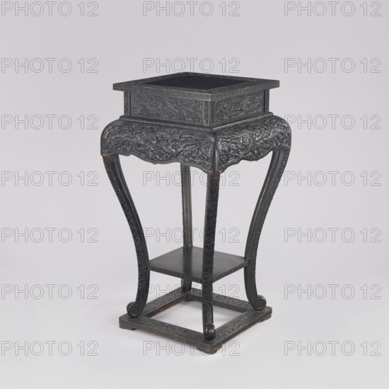 Table, phoenixes and dragons iconography, Chinese art, Chinese culture, Qing 1644–1911, 19th century, furniture, piece of furniture, height 77.0 cm, width 41.0 cm, depth 36.6 cm, A carved wooden stand with a square top and a smaller square upper compartment sits on a curved four-legged base; the wood appears dark with areas of worn brown showing through, and the surface is covered in intricate raised carvings of swirling patterns and stylized motifs. The legs curve outward and end in scroll-like feet, connecting to a square stretcher base that also has carved decoration; a small shelf is set between the legs below the upper section. The visible colors are dark brown, black, and hints of lighter brown where the finish is worn.