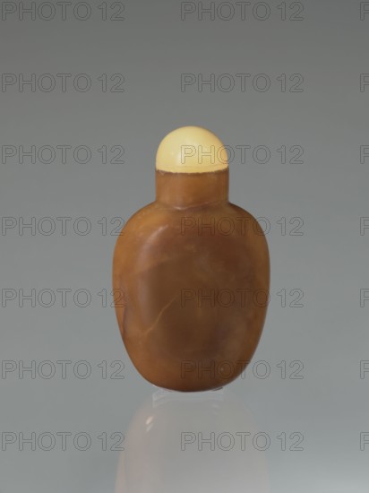 Snuffbox, gift; Chinese; Qing dynasty 1644–1911, 19th century; vessel; height 6.8 cm, width 4.0 cm, depth 2.4 cm., A smooth, rounded object resembling a small bottle or flask sits upright against a muted gray background, with a dome-shaped cap in a pale yellow color and a body in warm brown tones showing subtle swirls and lighter patches; the surface appears slightly glossy with soft reflections and a faint shadow and reflection beneath it on the surface it rests on.