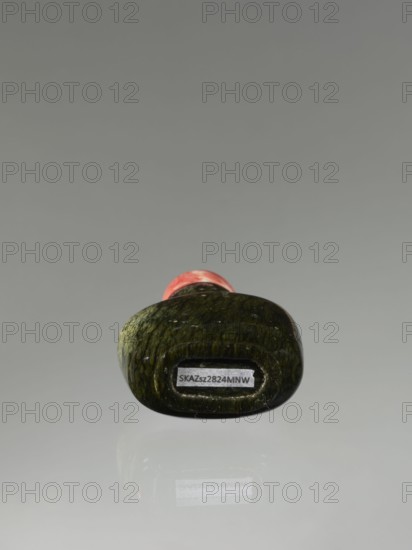 Snuffbox with spoon, Chinese, Qing dynasty 1644–1911, 19th century. Moon bottle snuffbox carved from rhodochrosite plug stone. Vessel; height 7.1 cm, width 4.6 cm, depth 2.6 cm., An object resembling a small rounded sculpture rests on a reflective surface, showing a textured dark green body with mottled lighter green highlights and a small visible area of pinkish red near its top; the background and reflection are shades of gray.