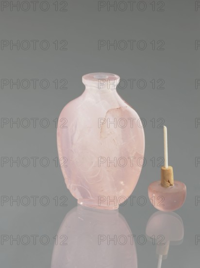 Snuffbox with spoon; maker, ; decorated with flowers and dragonflies; Chinese art; Chinese culture; Qing dynasty 1644–1911, 19th century; China; vessel; height 7.4 cm, width 4.4 cm, depth 2.5 cm., A translucent pink rounded bottle with a short neck and flared rim stands next to a small rounded stopper topped with a beige cylindrical piece and a thin pale stick; the bottle shows carved floral patterns and a visible chip on one side, both objects rest on a reflective surface and are set against a smooth gray background with pale pink and soft beige tones visible in the reflections.