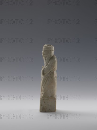 Sage, figurine; men; Chinese art; Chinese culture; Qing 1644–1911, 19th century; China; sculpture; height 16.8 cm, width 4.6 cm, depth 4.3 cm., A carved stone figure standing upright on a rectangular base, rendered in pale greenish-beige with faint areas of brownish veining and slight reddish stains, depicted in a long robe with flowing folds and a long beard, wearing a rounded hat and holding its hands together at the front, shown in profile against a smooth gradient background that transitions from dark gray at the top to light gray at the bottom.
