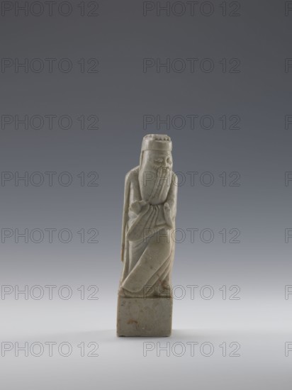 Sage, figurine, men, Chinese art, Chinese culture, Qing 1644–1911, 19th century, China, sculpture; height 16.8 cm, width 4.6 cm, depth 4.3 cm., A small carved stone figurine stands on a square base against a neutral gradient background; the figure has a long carved beard and mustache, a tall hat, and is robed with draped folds, showing a folded arm and a hand near the chest. The stone is mostly pale gray with subtle variations and a few small areas of brownish-orange spotting, and the background transitions from dark gray at the top to light gray and nearly white near the bottom.