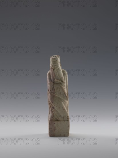 Sage with a Fan, figurine of a man holding a fan, Chinese art, Chinese culture, Qing dynasty 1644–1911, 19th century, China, sculpture; height 16.1 cm, width 4.4 cm, depth 4.3 cm, A carved stone figure standing on a rectangular base, depicted with a long robe and a tall headdress; the figure's hands are brought close to the face with one arm crossing the body, and the robe shows carved folds and drapery. The stone has a pale greenish-beige tone with scattered brownish spots and mottling, and the background is a smooth gradient of dark gray at the top fading to light gray near the bottom.