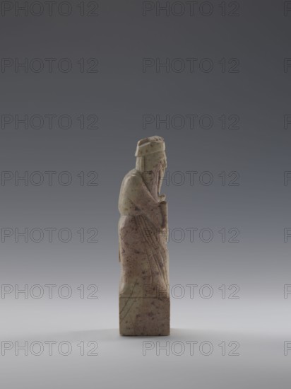 Sage with a Fan, figurine of a man holding a fan, Chinese art; materials: sculpture; culture: Chinese, Qing dynasty 1644–1911, 19th century; dimensions: height 16.1 cm, width 4.4 cm, depth 4.3 cm., A small carved figure standing on a rectangular base, shown in profile facing right, with a long robe that has incised folds and a long beard; the figure appears to wear a hat and holds an object close to its chest. The material of the carving is a pale greenish-beige with darker brown and purple speckling and veins. The background is a smooth gradient of gray tones transitioning from darker at the top to lighter near the bottom.