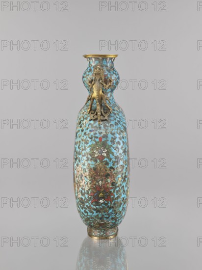 Vase — moon bottle pilgrim with a sleeve; maker, ; cloisonné enamel; Qing dynasty 1644–1911, 19th century, China; subject/iconography: peacocks, herons, dragons; ornament: flower tendril, lotus tendril; Chinese art/culture; vessel; height 39.3 cm, width 28.5 cm, depth 11.4 cm., A tall, narrow vase with a bulbous middle and a flared neck sits against a plain light gray background, the vase surface covered in an ornate pattern of turquoise, gold, dark brown, red, green, and white hues, and topped and rimmed with metallic gold; a decorative gold-colored sculpted ornament resembling a stylized animal motif is attached beneath the neck, and the vase casts a faint reflection on the glossy surface below.