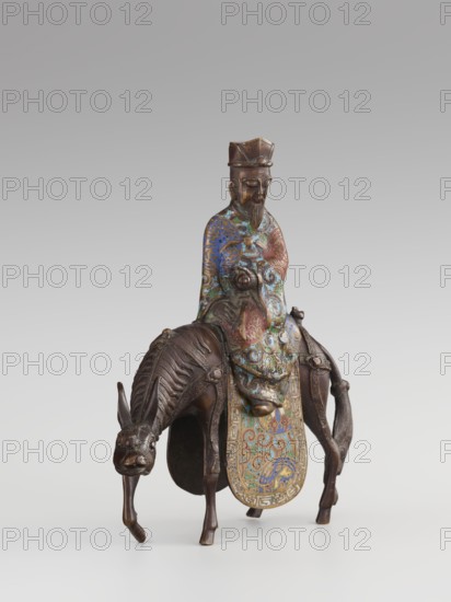Sage on a Mule;, cloisonné enamel; Qing 1644–1911, 19th century; China; sculpture depicting a sage riding a mule with men and figurines, mules, birds and zoomorphic motifs, plant motifs, meander ornament and scrolls; height 26.0 cm, width 25.5 cm, depth 6.0 cm., A small sculptural figure of a seated person wearing a tall hat and a long, patterned robe with visible swirling designs in blue, green, red, and gold tones, seated atop an animal resembling a horse or mule rendered in dark brown material with textured mane and limbs; the animal has a downward-tilted head and elongated ears, and there is a decorated cloth or saddle hanging down the animal's front with intricate multicolored patterns in blue, green, red, and gold; the figure has a detailed facial beard and a calm, closed-mouth expression, and the whole object rests on a neutral light-gray background.