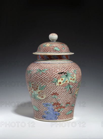 Vase with cover, 18th century, China; porcelain vessel with domed cover; Qing dynasty references to Ming/Wanli styles; ceramics decorated with fish scale ornament and pomegranates; backdated signatures; height 30.5 cm, diameter 19.5 cm; signed., A lidded ceramic jar with a rounded body and finial-topped lid stands on a light surface against a dark background. The jar is covered in a repeating scale-like pattern in red with areas of pale cream showing through, and it features painted floral and botanical motifs in green and pale yellow with touches of red. There are accents of blue and violet near the lower body and around the shoulder, and the lid has similar red scale patterning with green leaf-like shapes and a cream-colored knob with small red markings. The overall colors visible include red, cream, green, blue, pale yellow, violet, and black outlines.