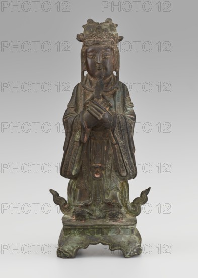 Dignitary — official; maker, figurine of a man; Chinese art; culture: Chinese; subject: scholars, sages, government officials; material: sculpture; date: 19th century Qing dynasty, 1644–1911; dimensions: height 28.5 cm, width 12.4 cm, depth 8.7 cm., A standing weathered statue with a patina of green, brown, and dark bronze tones depicts a robed figure wearing an ornate crown, holding a small object with both hands crossed at chest level; the face is serene with closed lips and finely carved facial features, and long earflaps or hair hang beside the face. The flowing robes display detailed carved patterns and layered sleeves with traces of darker brown and coppery highlights, and the figure stands on a square base that shows carved decorative elements and areas of green corrosion. Two upward-curving decorative elements extend from the base near the figure’s feet, and overall the surface shows aged texture, spots of verdigris green, dark brown, and hints of reddish-brown.