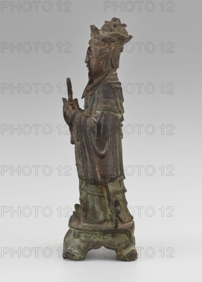 Dignitary - official, unknown,  craftsman author, figurine of a man, Chinese art, Chinese culture, Qing 1644–1911, 19th century, scholars, sages, government official, sculpture, height 28.5 cm, width 12.4 cm, depth 8.7 cm, A standing bronze figure with a greenish patina and areas of dark brown, shown in profile facing left, wearing a tall ornate headdress and layered robes with carved decorative patterns; the figure holds a small upright object in both hands near chest height, and the robes feature flowing sleeves and detailed borders; the base is a carved platform with a mottled green and brown surface and curved feet, and traces of wear and oxidation are visible across the statue.
