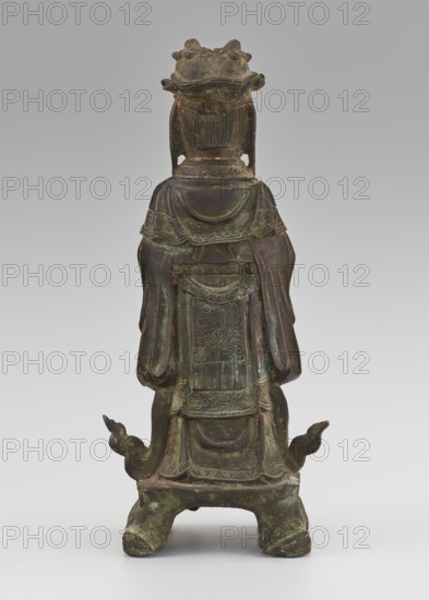Dignitary - official, unknown,  craftsman author, figurine of a man, Chinese art, Chinese culture, Qing 1644–1911, scholars, sages, government officials, 19th century, China, sculpture, height 28.5 cm, width 12.4 cm, depth 8.7 cm, A single standing bronze figure seen from the back, with a dark greenish-brown patina and areas of lighter green oxidation. The figure wears layered drapery with carved decorative borders and a rectangular panel on the back that has intricate low-relief patterns. The head is topped by a crown-like headdress with rounded projections, and the hair or headpiece has vertical striations. Sleeves hang down on either side and the hem of the garment flares slightly above a shaped base. The base rests on four short, curved feet with curled extensions at the sides, and the surface shows uneven texture and traces of wear in shades of brown, green, and hints of darker tones.