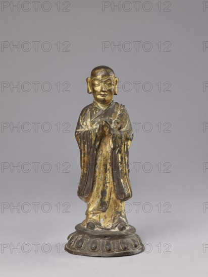 Monk on a Lotus Throne, lotus throne iconography, figurine, men, Chinese art, Chinese culture, Qing 1644–1911, Luohan iconography, arhat iconography, Chinese Buddhism, Buddhist art, 19th century, China, sculpture; height 31.0 cm, width 13.5 cm, depth 12.7 cm, A small standing figure with a rounded head and pronounced ears, wearing layered robes and holding hands together near chest level, positioned on a rounded ornate base; the surface shows worn gold and dark brown areas with patches of lighter and darker tones, and the background is a smooth gray.
