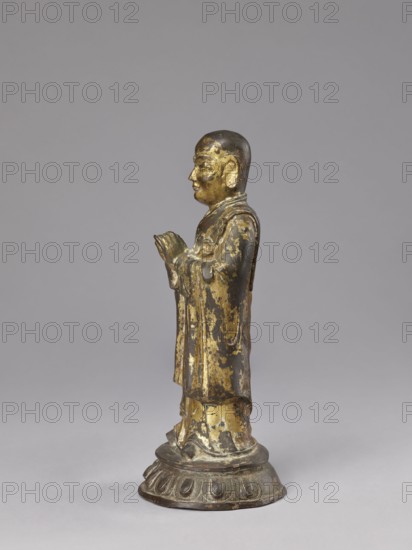 Monk on a Lotus Throne, lotus throne iconography, figurine, men, Chinese art, Chinese culture, Qing 1644–1911, Luohan iconography, arhat iconography, Chinese Buddhism, Buddhist art, 19th century, China, sculpture, height 31.0 cm, width 13.5 cm, depth 12.7 cm, A small standing figure rendered in dark metal with worn gold-colored gilding, shown in profile facing left with hands held together near the chest; the figure wears flowing layered robes that display patches of gold and darker exposed metal, and stands on a rounded base with petal-like relief details that are also dark with traces of gold.