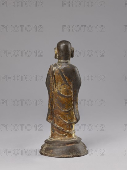 Monk on a Lotus Throne, lotus throne iconography, figurine, men, Chinese art, Chinese culture, Qing 1644–1911, Luohan arhat iconography, Chinese Buddhism, Buddhist art, 19th century, China, sculpture; height 31.0 cm, width 13.5 cm, depth 12.7 cm, A standing statue shown from the back, depicting a robed figure on a rounded base; the figure has a smooth, rounded head with pronounced ears and draped garments with visible folds. The surface shows worn areas and flaking, revealing a mix of dark brown and black tones with patches of golden yellow and muted ochre, and a few lighter beige spots on the base and robe. The background is a uniform soft gray.