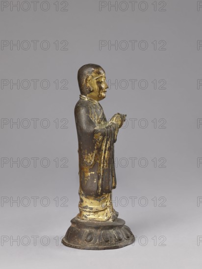Monk on a Lotus Throne, lotus throne iconography, figurine, men, Chinese art, Chinese culture, Qing 1644–1911, Luohan arhat iconography, Chinese Buddhism, Buddhist art, 19th century, China, sculpture; height 31.0 cm, width 13.5 cm, depth 12.7 cm, A small standing figurine viewed in profile on a rounded base, the figure wearing a long robe with draped sleeves and hands held together in front; the surface shows worn gilding and exposed darker underlying material, with visible areas of gold and brown, and the base has a darker brown tone with lighter worn spots; the background is a uniform light gray.