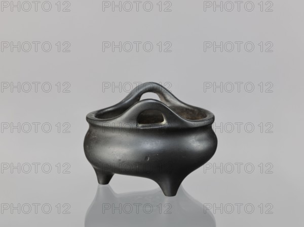 Li-type incense burner, ; decorated with cicadas, lion, and dragon iconography; Chinese, Ming dynasty Xuande period, 1426–1435, possibly 1426–1435; ceremonial/ritual bronze vessel; height 8.1 cm, diameter 11.1 cm., A small rounded vessel with a smooth matte surface sits on a reflective surface; it has a bulbous body, a flared rim with integrated curved handles that form arches, and stands on three short tapered legs. The vessel is primarily black with subtle variations in tone, and the background is a soft light gray.