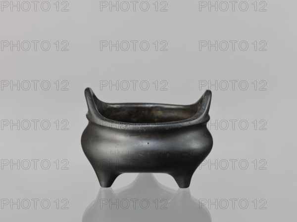 Li-type incense burner, cicadas and lion iconography, dragons, Chinese art/culture, Ming dynasty 1368–1644, Xuande period 1426–1435, ceremonial/ritual vessel, China, vessel, height 8.1 cm, diameter 11.1 cm, A small rounded vessel with a dark, almost black surface sits centered on a smooth, reflective surface; it has a wide, open top with two upward-curving rim extensions on opposite sides, a bulging body that narrows toward the base, and three short, tapered legs; the interior appears darker and slightly worn, and the surrounding background is a soft neutral gray.
