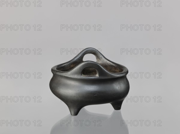 Li-type incense burner, cicadas, lion iconography, dragons iconography, Chinese art, Chinese culture, Ming 1368–1644, Xuande 1426–1435, 1426–1435, China, ceremonial/ritual vessel, height 8.1 cm, diameter 11.1 cm, A small, rounded three-legged vessel with a smooth, glossy black surface and a slightly reflective sheen sits on a pale gray surface against a light gray background, featuring a curved rim with two arched handle openings and an interior that appears darker with subtle brownish discoloration.
