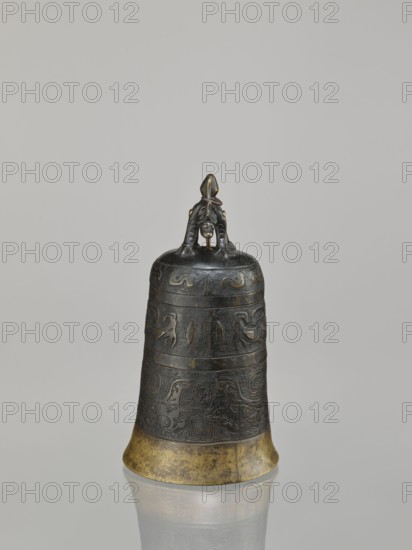 Bell, dragons iconography, Chinese art, Chinese culture, Qing 1644–1911, dragon with pearl, taotie iconography, meander ornament, leiwen ornament, 18th century, China, height 28.0 cm, width 15.3 cm, weight 2.29 kg, A dark metallic bell with a rounded top and a flared base stands upright on a reflective surface, showing a reflection beneath it. The bell has an ornate handle formed by a sculpted figure in a crouching position, with a small suspended element beneath the figure. The body of the bell is decorated with carved or cast patterns in horizontal bands, featuring raised motifs and textured designs. The lower rim of the bell has a worn, golden-colored band contrasting with the darker patina of the upper sections. Visible colors include dark bronze, black, muted gold, and subtle greenish-brown tones on the metal, set against a light gray background.