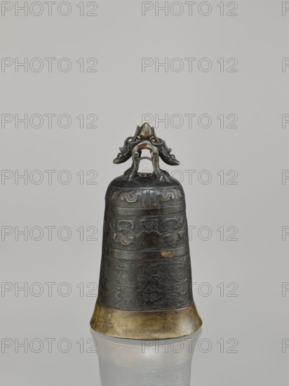 Bell, dragons iconography, Chinese art, Chinese culture, Qing 1644–1911, dragon with pearl, taotie iconography, meander ornament, leiwen ornament, 18th century, China, varia, height 28.0 cm, width 15.3 cm, weight 2.29 kg, A bronze bell with a dark patina and areas of golden bronze is shown standing upright on a reflective surface; the bell has carved decorative bands with swirling, stylized motifs around its body and a flared golden rim at the base, and its top features an ornate handle resembling intertwined animal forms with curved limbs and rounded heads in darker bronze with golden highlights, all set against a plain light gray background.