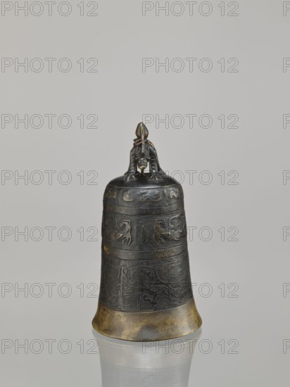 Bell, dragons iconography, Chinese art, Chinese culture, Qing 1644–1911, dragon with pearl, taotie iconography, meander ornament, leiwen ornament, 18th century, China, varia, height 28.0 cm, width 15.3 cm, weight 2.29 kg, A dark bronze bell with a slightly flared base sits upright on a reflective surface, showing a muted reflection beneath it; the bell features a carved banded design around its body with ornamental raised patterns and a weathered patina that reveals areas of brownish gold, dark green, and black; the top of the bell has a sculpted handle in the form of a small crouching figure in the same dark bronze tones, and the background is a smooth pale gray.