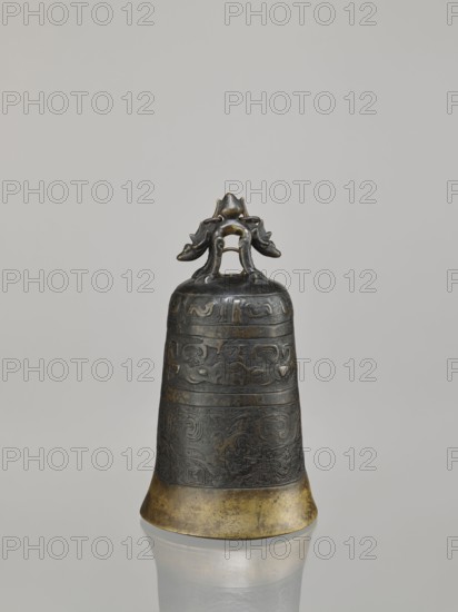 Bell, ; dragons iconography; Chinese art; Chinese culture; Qing dynasty 1644–1911; dragon with pearl; taotie iconography; meander ornament; leiwen ornament; 18th century; China place of origin; height 28.0 cm; width 15.3 cm; weight 2.29 kg, A single metal bell with a dark aged surface and a golden lower rim stands upright against a plain light gray background, featuring an ornate handle shaped like a stylized creature with curled limbs and decorative motifs; the bell body shows banded raised patterns and intricate carved designs in darker and slightly greenish-brown tones, and the base reflects faintly on the smooth surface beneath.