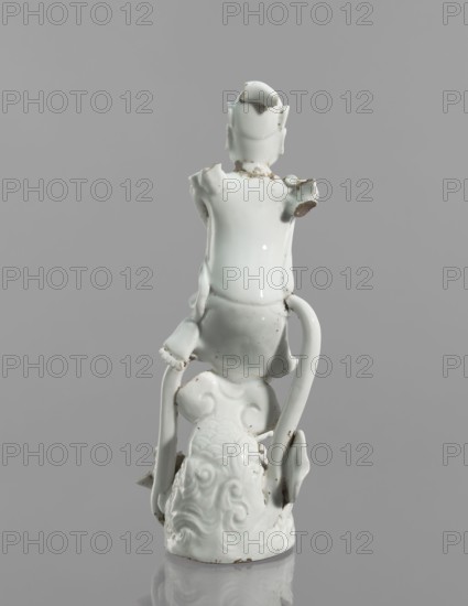 Kui Xing figurine with a leg resting on a mythical fish-dragon; ceramics; Chinese; 18th century; sculpture; height 29 cm; iconography: Kui Xing, dragons, ao mythology., A white glazed ceramic figurine with a glossy finish is shown from the back against a gray background; the figure is seated on an ornate base with swirling decorative relief and has elongated curved elements extending downward on both sides; the surface shows chipped and worn areas with exposed darker material at some edges and joints, and there are small patches of discoloration on the glaze.