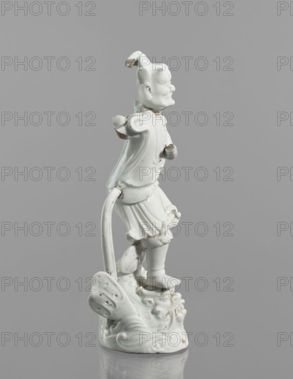 Kui Xing figurine with a leg resting on a mythical fish-dragon; ceramics; Chinese; 18th century; sculpture; height 29 cm; iconography: Kui Xing, dragons, ao mythology., A white ceramic figurine of a person standing on a decorative base, shown in profile against a plain gray background; the figure wears flowing garments and a skirt-like lower piece, has an upturned face with defined facial features and a hair or headpiece element, and one arm extended while the other is bent; the base includes stylized wave and shell-like forms, and there are small areas of surface discoloration and roughness on parts of the figure and base; visible colors are white, gray, and small brownish spots.