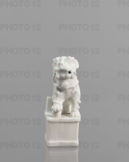 Foo lion figurine with a ball;, lion iconography; ceramics; blanc de Chine porcelain; figurine; Chinese art/culture; 18th century China; sculpture; height 13.2 cm, width 4.8 cm, depth 6.7 cm., A glossy white ceramic figurine of a stylized animal sits on a rectangular base against a smooth gray background. The animal has a rounded head with pronounced facial features, a wide open mouth showing teeth, and textured details on the forehead and ears. A collar with a rounded decorative element encircles the neck. The body shows stylized limbs with defined paws and small raised bumps along the sides. The rectangular base has smooth edges and a small cylindrical protrusion at one corner. Visible colors are white for the figurine and gray for the background.