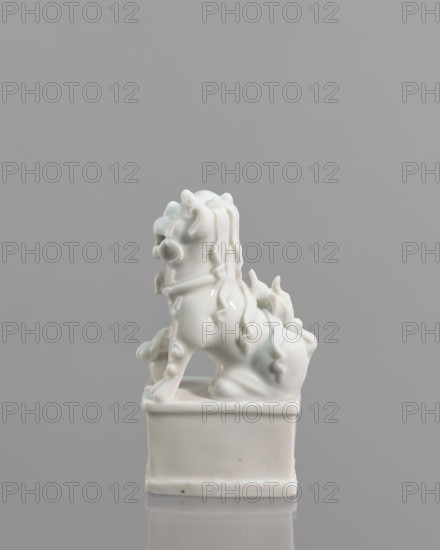 Fo lion foo dog figurine with a ball; maker, 18th century China; blanc de Chine porcelain; subject: lion foo dog iconography; form: animal sculpture/figurine; dimensions: height 13.2 cm, width 4.8 cm, depth 6.7 cm., A glossy white ceramic sculpture of a stylized mythical lion-like creature sits atop a rectangular pedestal, showing flowing, curled mane and tail details with smooth, rounded limbs and a slightly open mouth; the sculpture and pedestal rest on a reflective surface, and the background is a uniform gray. The visible colors are white, gray, and subtle pale greenish-blue tints on parts of the glaze.