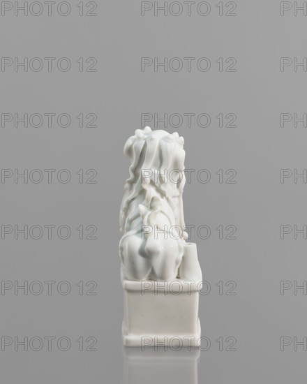 Fo lion figurine with a ball;, lion iconography; ceramics; blanc de Chine porcelain; figurine; Chinese art/culture; animal figurine; 18th century, China; sculpture; height 13.2 cm, width 4.8 cm, depth 6.7 cm., A glossy white ceramic figurine sits on a rectangular base against a neutral gray background. The figurine features flowing, wavy forms that cascade down the back, creating a sense of layered, ribbon-like texture. The base and the figure share the same smooth, reflective white surface, with subtle highlights and soft shadows emphasizing the curves and folds. The overall composition is centered in the frame, with the gray background appearing uniform and uninterrupted. Colors visible are white and gray.