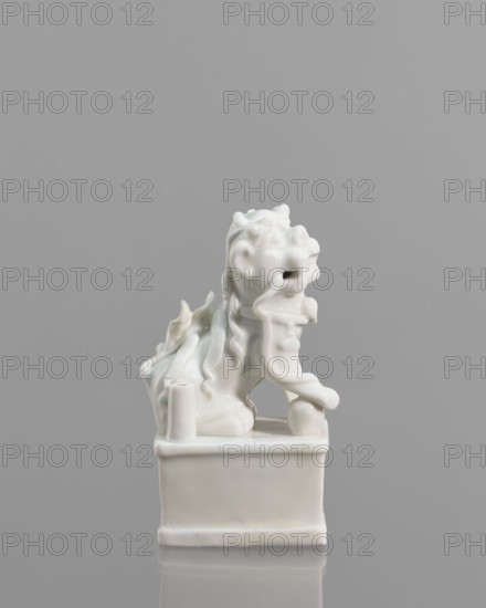 Fo lion figurine with a ball;, lion iconography; blanc de Chine porcelain; animal figurine; Chinese art; 18th century, China; sculpture; height 13.2 cm, width 4.8 cm, depth 6.7 cm., A glossy white ceramic figurine of a stylized lion-like creature sitting on a rectangular plinth, shown against a solid gray background; the creature has a flowing mane with wavy, ribbon-like strands, a pronounced open mouth, curled features on its head, and a raised forepaw resting on a spherical object, with a few curved ornamental elements along its back and sides.