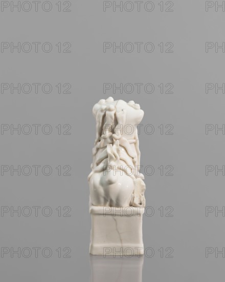 Incense burner in the shape of a fo lion with a ball; ceramic sculpture; Chinese; 18th century; height 13.0 cm, width 6.0 cm, depth 4.8 cm; censers/incense stand; animal figurine; lion iconography; fo iconography., A small glossy white sculptural object sits on a rectangular base against a plain gray background; the object has a smooth, rounded lower section and an intricately detailed upper portion with flowing, ribbonlike forms and bulbous knots that cascade downward, creating a layered, draped appearance, and there are subtle hairline cracks and slight discolorations visible in the white surface.