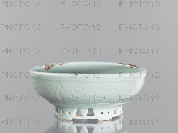 Bowl on an openwork foot. Chinese, Qing dynasty, Yongzheng period 1723–1735. Colored porcelain decorative vessel featuring fish carps and crabs. Height 9.5 cm; diameter 23.8 cm., A shallow ceramic bowl with a pale celadon glaze sits on a reflective surface; the bowl has an openwork foot with rounded cutouts and a slightly flared rim, the exterior surface showing subtle raised decorative patterns and a few small darker specks, while the interior rim features small floral motifs in reddish tones; visible colors include pale green, light blue-green, reddish accents, white highlights, and the gray background and reflection.