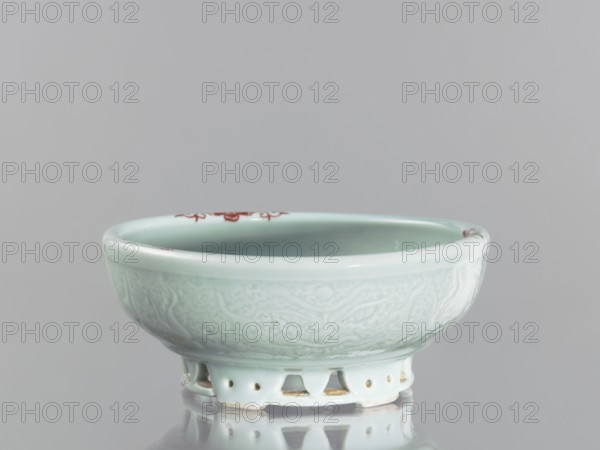 Bowl on an openwork foot, Chinese, Qing dynasty, Yongzheng period 1723–1735. Porcelain with polychrome decoration of fish carps and crabs. Vessel, height 9.5 cm, diameter 23.8 cm. China., A shallow bowl with a pale celadon glaze sits on a short openwork foot, showing a smooth glossy interior and an exterior with subtle raised decorative patterns; the rim has small areas of reddish-brown detail. The visible colors are pale green, white, reddish-brown, and soft gray from the background and reflection.