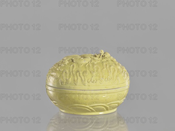 Box with a lid, Kangxi style, ceramics, Qing style, dragons iconography, Chinese art, Chinese culture, pearls, clouds, Qing 1644–1911, Kangxi 1662–1722, 18th century, China, vessel; height 5 cm, diameter 7.4 cm., A small round lidded object sits centered on a reflective surface, the piece is a pale yellow color with a glossy finish and subtle speckling, its lid features raised swirling and wave-like decorative patterns and a sculpted motif on top, the body below the lid has carved curved lines and banding around its midsection and base, the background and reflection are shades of gray.