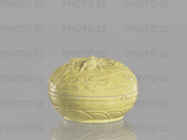 Box with a lid, Kangxi style, ceramics, Qing dynasty 1644–1911, Kangxi period 1662–1722, 18th century China; decorated with dragons, pearls and clouds; vessel; height 5 cm, diameter 7.4 cm., A small rounded lidded container sits centered against a plain neutral gray background, the object and its reflection visible on a glossy surface. The container is pale yellow with a glossy finish and shows carved or molded swirling patterns and wave-like motifs across its curved surfaces; the lid has a raised decorative element with textured details. The yellow surface has small darker speckles and a few tiny imperfections. The overall scene includes shades of yellow, gray, and subtle reflections creating slightly lighter and darker tones.