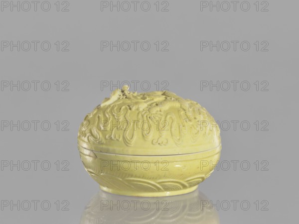 Box with a lid, Kangxi style, ceramics, Qing 1644–1911, Kangxi period 1662–1722, 18th century China; decorated with dragons, pearls, and clouds; vessel; height 5 cm, diameter 7.4 cm., A small round object with a lid sits centered against a neutral gray background, the object having a glossy pale yellow surface with a carved, raised textured pattern covering its upper half and a smoother lower half with incised curved motifs; the lid area shows sculpted flowing, looping relief and a small raised element at the top, while the base rests on a reflective surface that mirrors the object's yellow color and shapes.