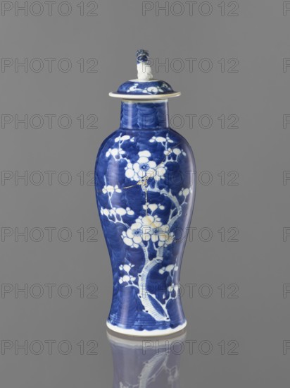 Meiping vase with cover, maker, ceramics; meiping vessel with cover; blue-and-white porcelain depicting plum trees and floral tendrils; Chinese art, China; 17th–18th century; height 31 cm., A tall blue and white ceramic vessel with a lid stands against a gray background, showing a glossy blue surface decorated with white floral and branch motifs; the lid has a small sculpted handle and a white rim, and the body displays visible hairline repairs highlighted by thin metallic lines; the base edge is white and the vessel casts a faint reflection below.