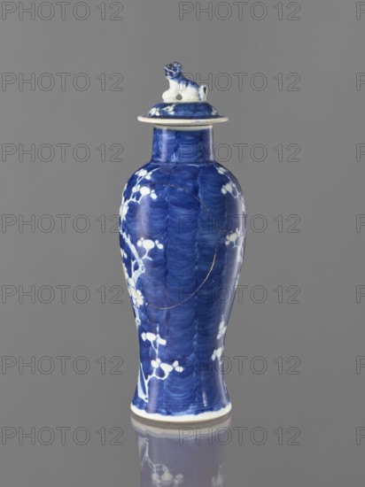 Meiping vase with cover, meiping vessel type, cover, blue and white porcelain, China, 17th–18th century, height 31 cm; decorated with plum trees and floral tendril motifs; ceramics;, A tall, slender ceramic vase with a lid sits against a neutral gray background; the vase is predominantly deep blue with areas of lighter blue brushlike textures and white floral or branch-like motifs scattered over its surface, and the lid features a small sculpted animal figure colored in blue and white; the base edge shows a thin white rim and the vase and its lid cast a subtle reflection on the glossy surface beneath.