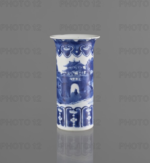 Flute-type vase, Chinese blue-and-white porcelain, landscape motifs; Kangxi period 1662–1722, 17th–18th century; China; vessel; height 15 cm, diameter 8.1 cm., A cylindrical ceramic vessel stands upright against a gray background, decorated with blue-painted scenes and patterns on a white base. The upper rim flares outward slightly and features a band of repeating heart-shaped motifs in deep blue. Below this, a painted scene shows architectural forms resembling a multi-tiered gate and surrounding structures, rendered in varying shades of blue with brushstroke textures and a small human silhouette visible near an archway. The lower section has a band of repeating vertical panels with floral or geometric motifs in dark and medium blue. The vessel casts a subtle reflection on the glossy surface beneath it. Colors visible include blue, white, and gray.