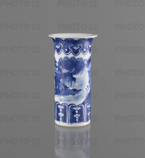 Flute-type vase, ceramics, Chinese blue-and-white porcelain with landscape motifs, Chinese, Kangxi 1662–1722, 17th–18th century, China; vessel; height 15 cm, diameter 8.1 cm., A tall cylindrical porcelain vase stands upright against a plain gray background, displaying a painted blue-and-white design; the rim flares outward slightly and is white, while a band of deep blue with lighter blue decorative motifs circles near the top; the main body shows a scene in varying shades of blue with brushwork suggesting curved architectural forms and flowing water, and a band of darker blue panels with lighter decorative elements wraps near the base; the vase sits on a reflective surface that mirrors its blue-and-white colors.