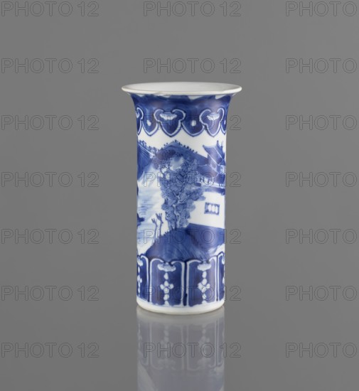 Flute-type vase, blue and white porcelain, landscape motifs, Chinese, Kangxi 1662–1722, 17th/18th century, China; vessel; height 15 cm, diameter 8.1 cm., A tall cylindrical ceramic vase stands upright against a smooth gray background, decorated in blue and white. The rim flares outward slightly and is white on top with a band of deep blue beneath, featuring a repeating scalloped motif. The central body shows a painted scene in blue tones with a landscape including a tree, hills, and architectural forms. The lower section has a decorative border in dark blue with vertical panels and small floral-like elements. The vase sits on a reflective surface that mirrors the blue and white shapes and the gray tones of the surroundings.