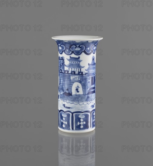 Flute-type vase, blue-and-white porcelain, Chinese, 17th–18th century, height 15 cm, diameter 8.1 cm; decoration: landscape motifs., A cylindrical ceramic vessel with a flared rim stands upright against a plain gray background; the vessel is primarily white with blue painted decorations depicting architectural shapes and decorative borders, including a darker blue scalloped band near the top, a scene of pagoda-like structures and a walled gate around the middle, and a patterned border with floral motifs near the base; the surface shows glossy reflections and a faint mirror-like reflection of the vessel appears on the gray surface below.