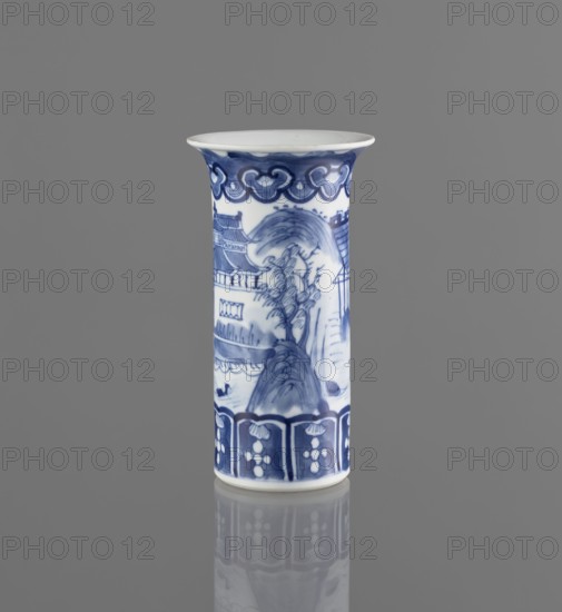 Flute-type vase, ceramic blue-and-white porcelain, Chinese, 17th–18th century, landscape motifs, vessel, height 15 cm, diameter 8.1 cm., A tall cylindrical ceramic vase stands upright against a plain gray background, decorated in blue and white with a glossy finish and a flared rim. The surface shows a painted landscape scene in various shades of blue, including stylized trees, a hill, and architectural elements, with a decorative band near the top featuring curved motifs and a patterned border near the base with floral-like shapes. The vase casts a faint reflection on the smooth gray surface beneath it, and the visible colors are blue, white, and gray.